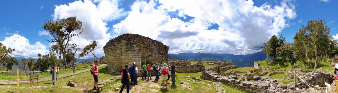 Panoramic view of ancient stone fortress ruins at Kuelap archaeological site in Peru with tourists walking among curved limestone walls under blue sky with white clouds and mountain landscape in background