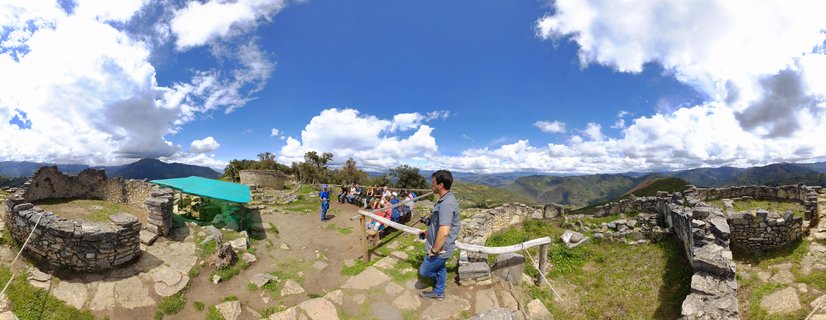 Panoramic view of ancient stone ruins at Kuelap archaeological site with tourists walking among circular stone walls, mountains and blue sky with clouds in background