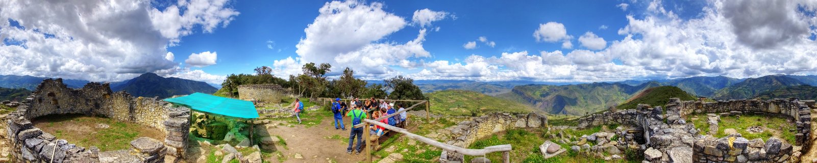 Panoramic view of ancient stone ruins at Kuelap archaeological site with tourists walking among stone walls, mountains and cloudy blue sky in background