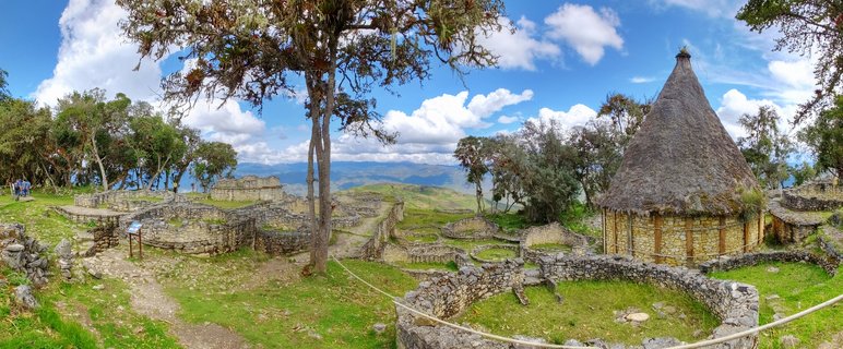 Ancient stone ruins of Kuelap fortress in Peru with circular foundations, reconstructed thatched-roof building, green grass, trees, and mountain views under blue sky with clouds