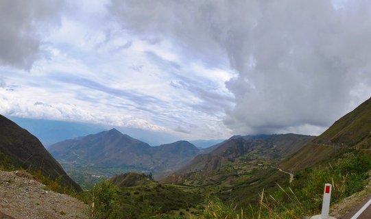 Mountain valley landscape showing green Andean hillsides with a winding road, small settlements in the valley floor, and dramatic cloudy sky over Cajamarca, Peru