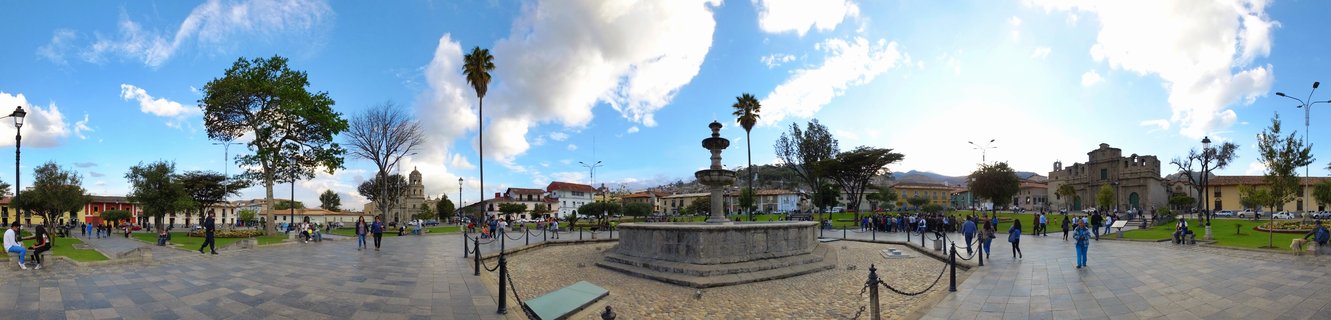 Wide panoramic view of a colonial town square with a stone fountain in the center, surrounded by historic buildings, palm trees, and people walking on paved pathways under a blue sky with clouds