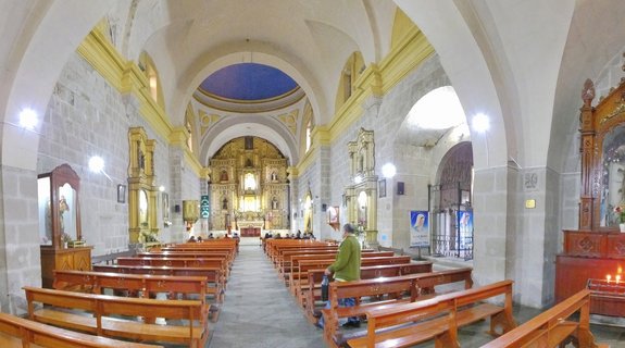 Interior view of a colonial church showing rows of wooden pews, stone arches with yellow painted trim, and an ornate golden altar at the far end under a blue dome