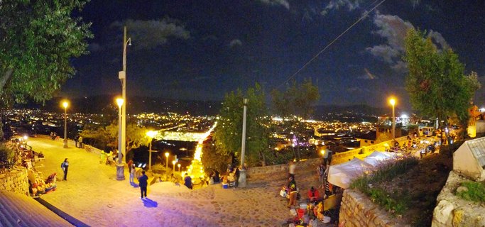 Panoramic night photograph showing an illuminated city spread across a valley, viewed from an elevated cobblestone terrace with street lamps, trees, and people in the foreground under a dark sky with scattered clouds.