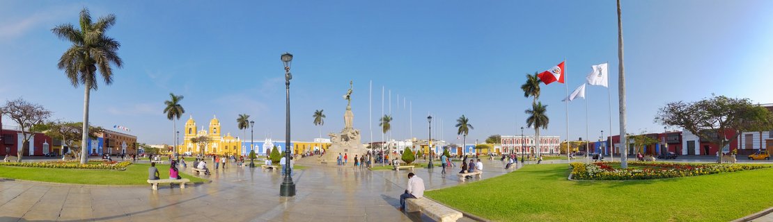 Wide panoramic view of a colonial plaza with a yellow cathedral, central monument statue, palm trees, green lawns, people on benches, and Peruvian flags against blue sky