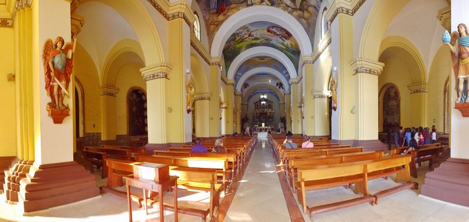 Interior view of a cathedral with yellow arched columns, wooden pews, religious angel statues, ceiling frescoes, and visitors sitting and walking toward the altar