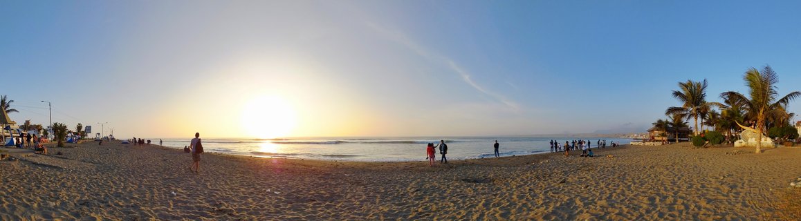 Wide beach panorama at sunset showing people walking on sand, ocean waves, palm trees on the right, and the sun setting over the Pacific Ocean