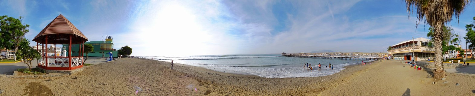 Panoramic beach scene showing a wooden gazebo with brown roof on sandy shore, people walking and swimming in ocean waves, a long pier extending into the water, beachfront buildings, and palm trees under blue sky with scattered clouds