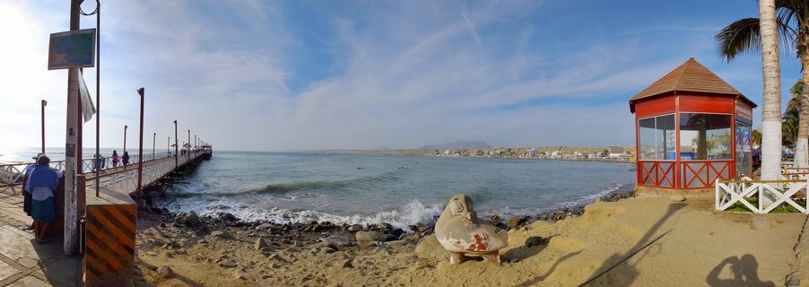 Coastal scene showing a wooden pier extending into ocean waters, a red gazebo on sandy beach, palm trees, and surfers in the water at Huanchaco, Peru
