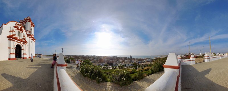Panoramic view from a white colonial church terrace with red trim overlooking Huanchaco town and Pacific Ocean coastline under blue sky with visitors on the walkway