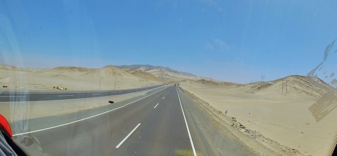 A paved highway running through desert landscape with sandy hills, mountains in the distance, and clear blue sky overhead
