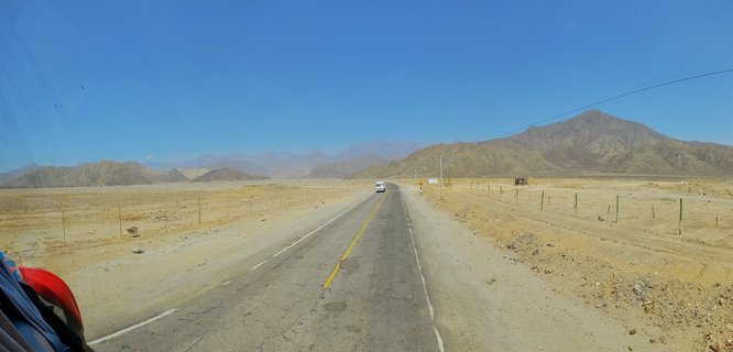 A panoramic view of a paved two-lane road with yellow center line running through desert landscape toward distant mountains under clear blue sky in Peru