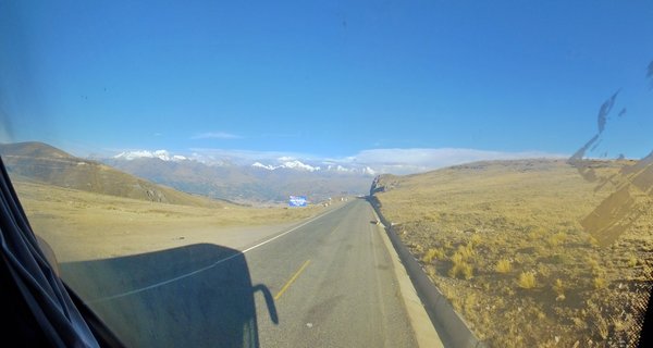 View from a vehicle window showing a highway through dry grassland with snow-capped Andes mountains in the background under clear blue sky