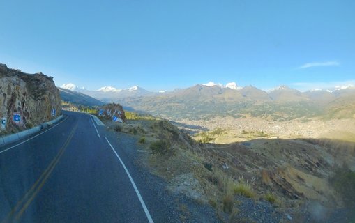 Panoramic mountain landscape showing snow-capped Andes peaks above Huaraz city in a valley, with a paved road and white bollards in the foreground and people sitting near a small wooden structure on the hillside