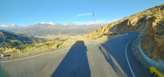 A winding mountain road with double yellow lines curves through dry golden hillsides toward snow-capped Andes mountains under clear blue sky, with a yellow winding road warning sign visible on the right