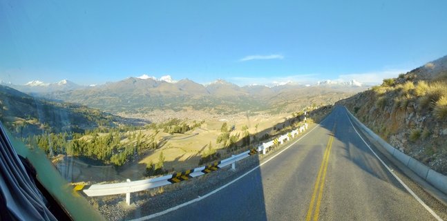 Winding asphalt mountain road with yellow double center lines and white guardrails ascending through dry grassland hills toward snow-capped Cordillera Blanca mountains, with Huaraz city visible in the valley below under clear blue sky