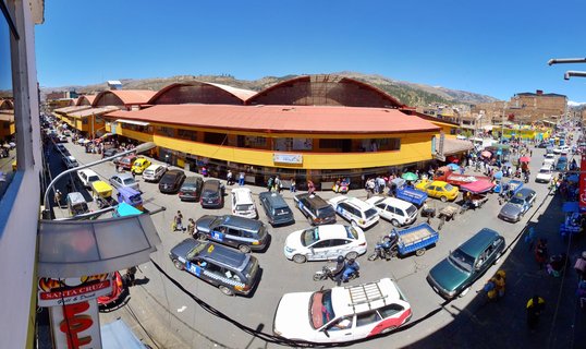 High-angle view of a busy street intersection in Huaraz, Peru, showing a large yellow market building with red arched roofs, numerous taxis and vehicles, pedestrians with umbrellas, and mountains in the background under a clear blue sky