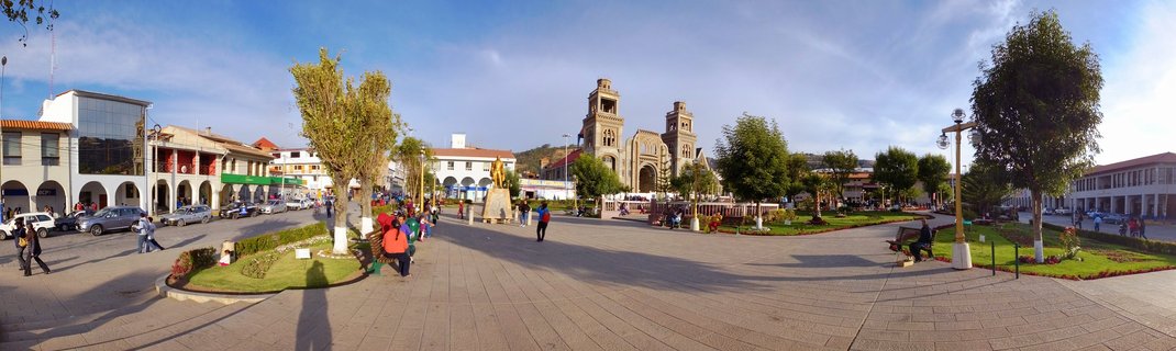 Wide panoramic view of Plaza de Armas in Huaraz, Peru showing the Cathedral with twin towers, paved plaza with pedestrians, trees, benches, and colonial buildings under blue sky