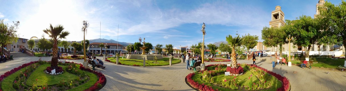 Wide view of a town plaza with colonial church, fountain, palm trees, flower gardens, people walking, and mountains in the background under blue sky