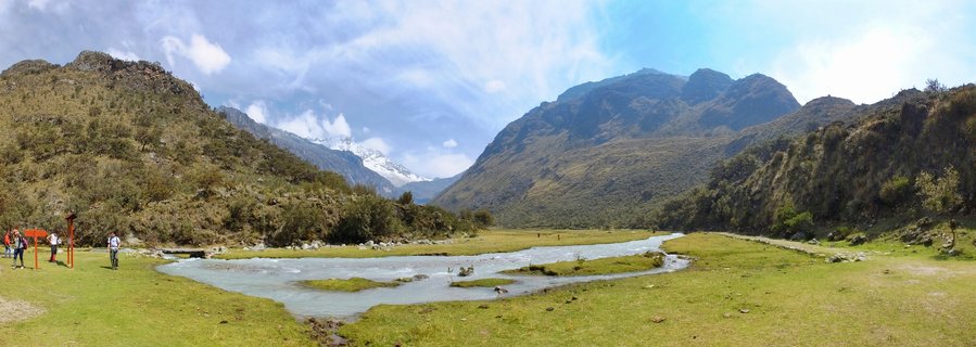 Panoramic landscape showing a mountain valley in Cordillera Blanca near Huaraz, Peru. A group of hikers stands on a grassy area near wooden signposts on the left. A winding milky-blue stream flows through green meadow in the foreground. Steep mountain slopes rise on both sides. Snow-capped peaks are visible in the distance under a partly cloudy sky.