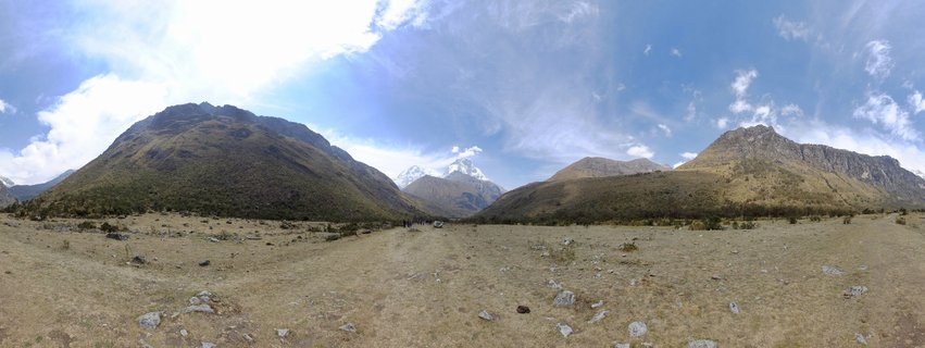 Wide mountain valley landscape showing snow-capped Andes peaks in the distance, brown grassy plains with scattered rocks in the foreground, and large mountain slopes on either side under a blue sky with white clouds