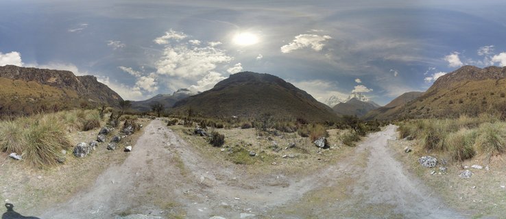 Panoramic landscape photograph showing a forked dirt road through high-altitude Andean grassland with mountains and snow-capped peaks in the background under a blue sky with scattered clouds