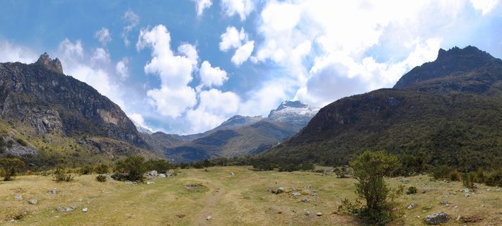 Mountain valley landscape showing snow-capped Andes peaks, grassy foreground with scattered shrubs, rocky slopes, and blue sky with white clouds near Huaraz, Peru