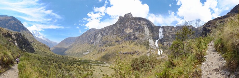 Panoramic mountain landscape showing hikers on a dirt trail through a green valley with a waterfall cascading down rocky cliffs, snow-capped peaks in the background, and blue sky with white clouds above the Andes mountains in Huaraz, Peru