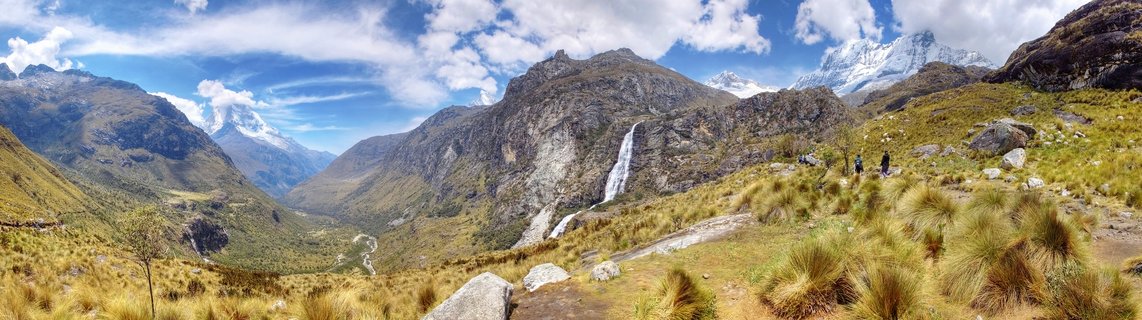 Panoramic mountain landscape showing snow-capped peaks of the Cordillera Blanca range in Peru, with a waterfall cascading down a rocky cliff, golden high-altitude grasses in the foreground, and small hikers visible on a trail under blue sky with scattered clouds