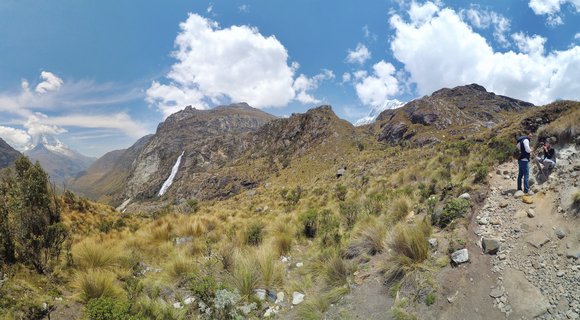 Two hikers on a rocky mountain trail in Peru's Huascarán National Park, surrounded by golden ichu grass with a waterfall cascading down the mountainside and snow-capped Andes peaks visible in the distance under a blue sky with white clouds