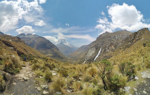 Mountain valley landscape in Huaraz Peru with stone hiking trail, golden grasses, distant snow-capped peak, waterfall, and blue sky with clouds