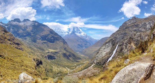 Snow-capped mountain peak centered in frame above a green-brown glacial valley with rocky slopes on both sides, a waterfall on the right mountainside, blue sky with white clouds above, large boulders in foreground with golden grass