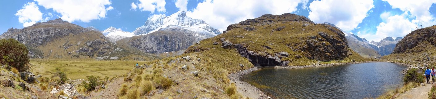 Panoramic view of snow-capped mountain peaks of the Cordillera Blanca range reflected in a turquoise alpine lake, with golden-brown grassy hillsides in the foreground and hikers visible on a trail, under a blue sky with white clouds near Huaraz, Peru