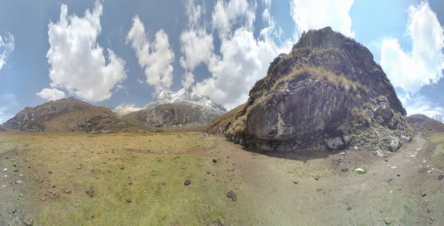 Panoramic landscape showing two hikers on a dirt trail through grassy alpine meadow with large rocky boulder formation on right and snow-covered mountain peaks in background under blue sky with white clouds