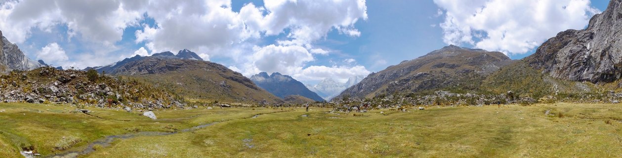 Wide mountain valley in Peru's Cordillera Blanca with rocky peaks, green-brown grassland, small stream, hikers in distance, and blue sky with white clouds