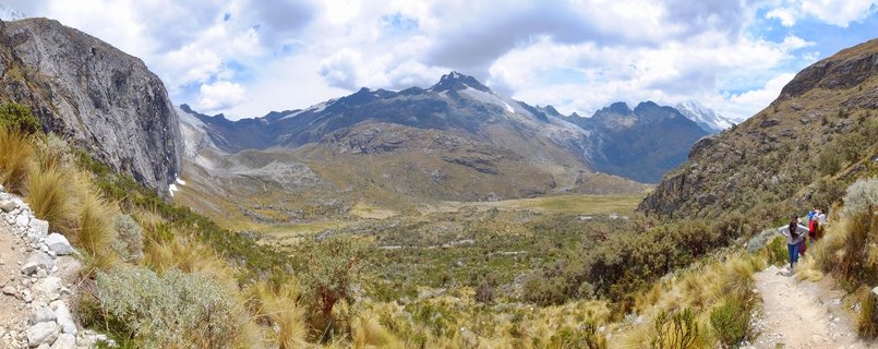 Panoramic landscape showing hikers on a mountain trail with snow-capped Andes peaks, rocky cliffs, and a wide alpine valley in Huaraz, Peru