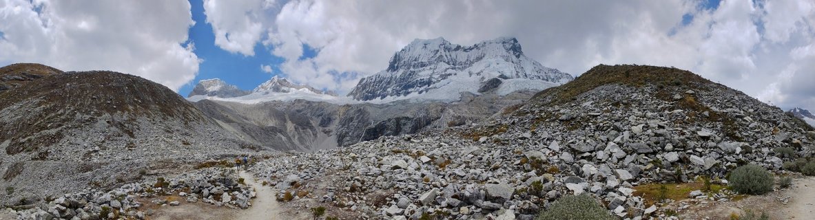 Panoramic view of snow-covered mountain peaks in Peru's Cordillera Blanca with rocky foreground terrain and hikers on a dirt trail under partly cloudy sky