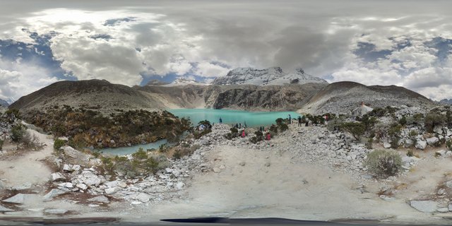 Group of hikers in outdoor gear walking along rocky shore of turquoise glacial lake with snow-capped Andes mountain peaks and cloudy sky in background near Huaraz, Peru