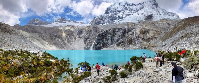 Panoramic view of turquoise glacial lake Laguna 69 surrounded by snow-capped mountains in the Peruvian Andes, with hikers visible along the rocky shoreline and alpine vegetation in the foreground