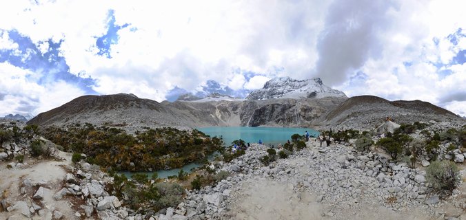 Panoramic landscape showing a bright turquoise alpine lake surrounded by rocky terrain and snow-capped mountain peaks under a cloudy sky, with small figures of hikers visible along the shoreline