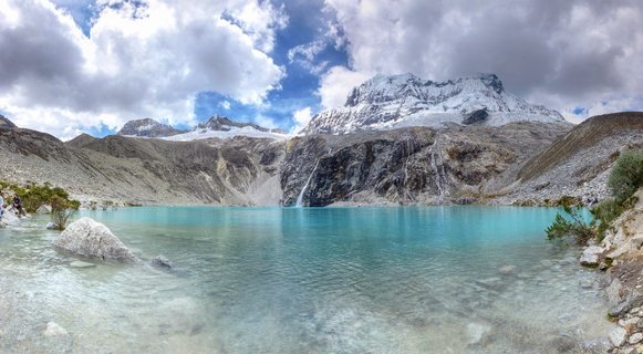 Panoramic view of a turquoise mountain lake with snow-covered peaks, rocky slopes, and a waterfall in the Peruvian Andes under a cloudy sky
