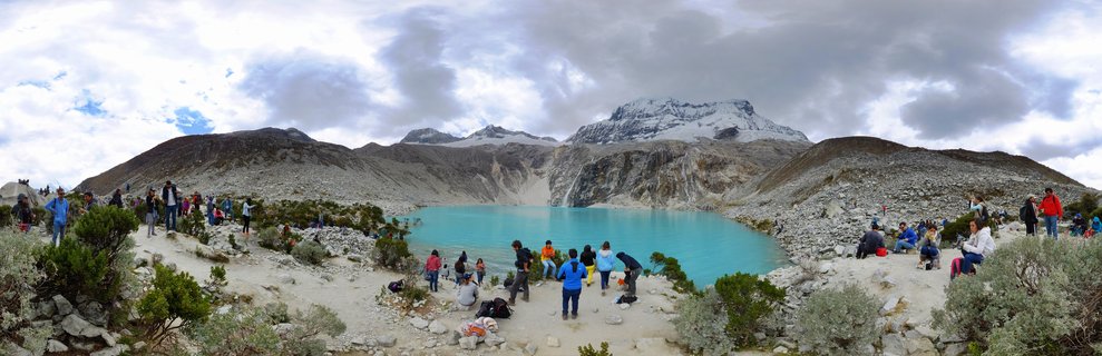 Wide panoramic view of tourists visiting a bright turquoise alpine lake surrounded by rocky mountains with snow-capped peaks in the Cordillera Blanca near Huaraz, Peru