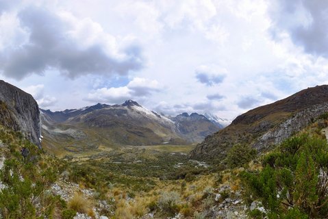 Wide mountain valley in the Peruvian Andes with snow-capped peaks in the distance, rocky slopes on either side, green and golden alpine vegetation in the foreground, and cloudy sky above