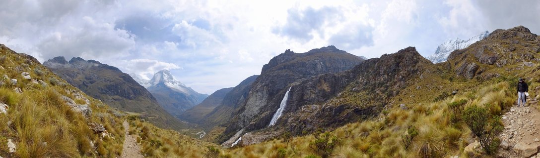 Panoramic mountain landscape showing a waterfall cascading down rocky cliffs in Peru's Cordillera Blanca, with snow-capped peaks, golden grassland, and a hiking trail with a hiker visible on the right