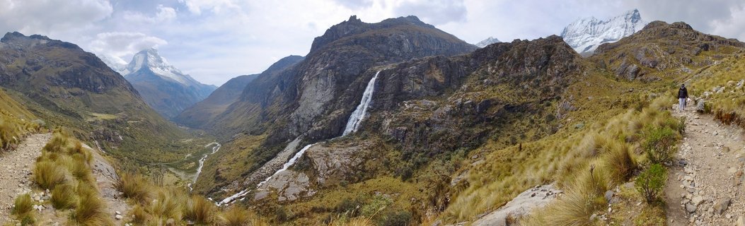 Panoramic view of a mountain valley in Peru showing a waterfall, snow-capped peaks, golden grassland, and a hiker on a dirt trail