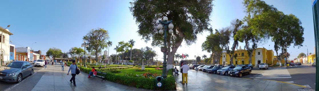 Wide street view of a Lima plaza with yellow colonial buildings, green park area with trees, pedestrians walking on sidewalks, and parked cars under clear blue sky