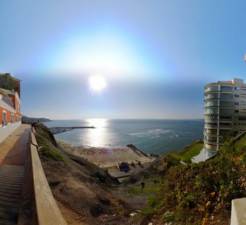 Aerial view from clifftop showing Pacific Ocean beach cove with people on sand, breakwater pier, modern white curved apartment building, orange building, cliff with erosion netting, and bright sun over water