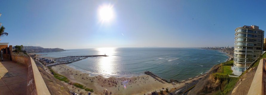 Panoramic coastal view from clifftop showing sandy beach with people, marina with boats, modern curved glass building, Pacific Ocean with sun reflection, and Lima cityscape along the coastline under clear blue sky