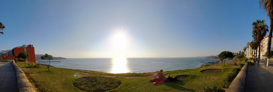 Wide coastal panorama showing the Pacific Ocean at sunset with green park grass in foreground, people sitting on blankets, palm trees, and apartment buildings along both sides
