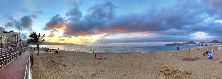 Wide panoramic photograph of Las Canteras beach at sunset showing golden sand, Atlantic Ocean waves, dramatic cloudy sky with orange and blue colors, palm trees, beachfront promenade with buildings, and people scattered across the beach